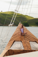 A woman in a lavender swimsuit sits cross-legged on the wooden bow of a sailboat, with water and a green hillside in the background.