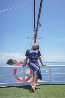 A woman in a navy blue two-piece outfit leans against a white railing on a boat deck, with the ocean, blue sky, and a pink life preserver in the background.