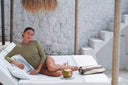 A woman reclines on a white outdoor daybed under a thatched roof, wearing a muted olive green long-sleeve dress