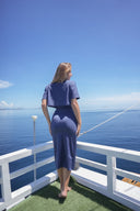A woman stands on a boat deck with a white railing, facing the open ocean under a clear blue sky. She is wearing a short-sleeve, high-neck, knee-length blue dress.