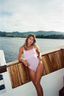 A woman wearing a light pink one-piece swimsuit leans against the wooden railing of a boat, with water and green hills in the background.