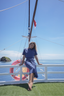 A woman wearing a matching blue short-sleeve top and skirt set sits barefoot on a white railing on a sailboat deck, with the blue ocean and sky in the background.