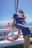 A woman in a blue short-sleeve dress leans back against a white railing on a boat deck, with a pink life preserver and blue sea and sky in the background.