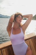 A woman wearing a light lavender one-piece swimsuit sits on a wooden boat railing with water and mountains in the background.
